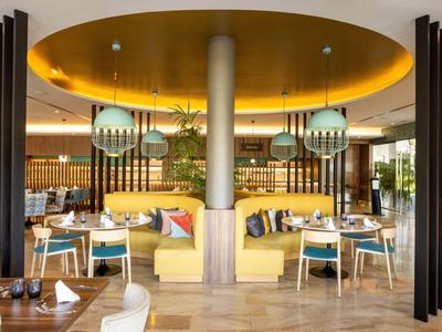 Modern restaurant met gele banken, ronde tafels en groene hanglampen.