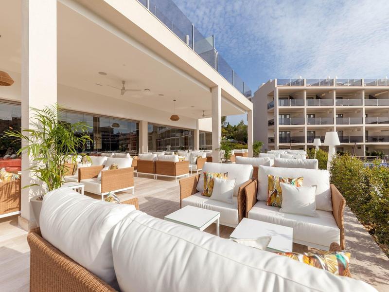 Grande terrasse avec mobilier de lounge blanc moderne devant un bâtiment hôtelier par temps ensoleillé.
