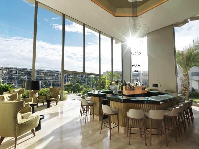 Bar d'hôtel lumineuse avec grandes fenêtres, sièges et vue sur la ville au coucher du soleil.