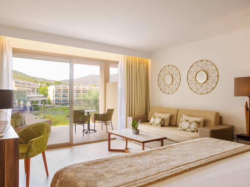 Chambre d'hôtel lumineuse avec lit double, coin salon et balcon donnant sur la piscine et le jardin.