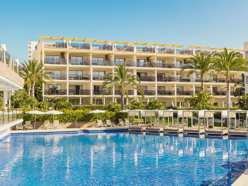 Hôtel à plusieurs étages avec balcons à côté d'une grande piscine avec chaises longues et palmiers.