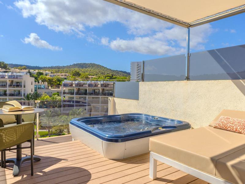 Terrasse avec bain à remous, chaise longue et vue sur les collines et bâtiments sous un ciel clair.