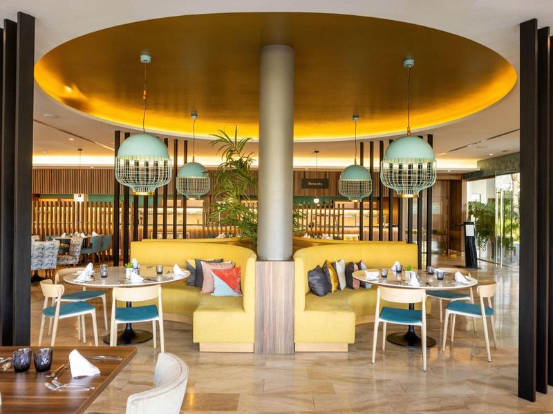 Salle à manger moderne avec banquettes jaunes, tables rondes et lampes suspendues.