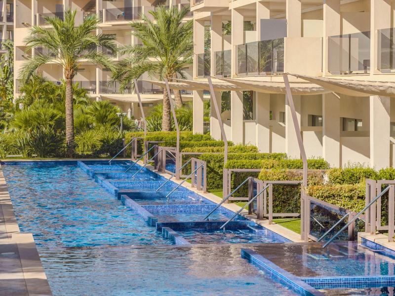 Piscine extérieure moderne avec carreaux bleus et chambres d'hôtel adjacentes, entourée de palmiers.