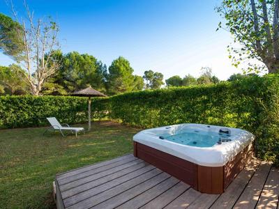 Buiten bubbelbad op houten terras met ligstoel en parasol, omringd door groene hagen en bomen.