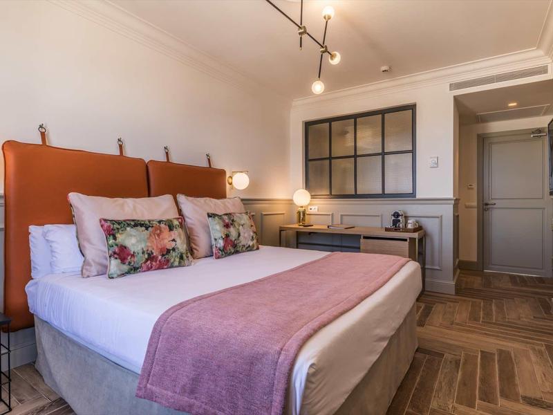 Modern ingerichte hotelkamer met groot bed, kleurrijke kussens en houten vloer.