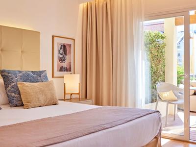 Světý pokoj v hotelu s pohodlnou postelí, jemnými závěsy a přístupem na terasu se židlemi.