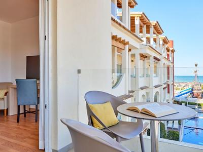 Balkon se židlemi a stolem, výhled na hotelovou budovu a moře v pozadí.