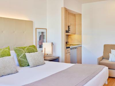 Světý hotelový pokoj s velkou postelí, zelenými polštáři, pohovkou a otevřeným kuchyňským koutem.