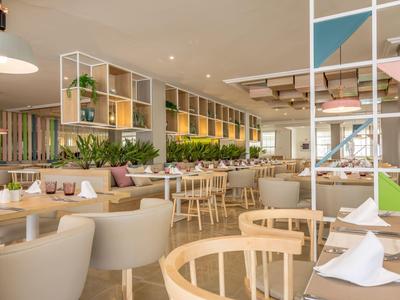 Světá moderní restaurace s dřevěným a bílým nábytkem a zelenými rostlinami.