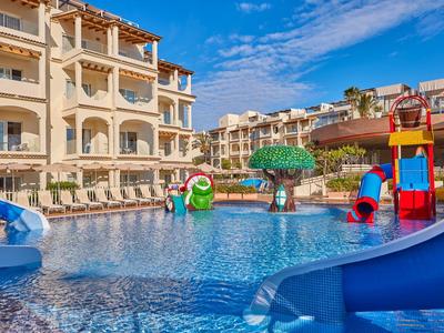 Dětský bazén s barevnými tobogány a herními prvky před hotelovou budovou pod modrou oblohou.