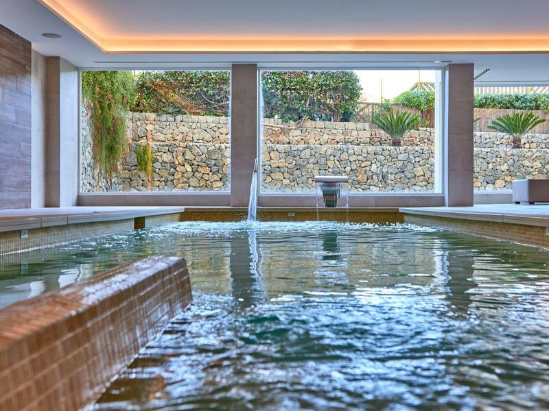 Moderní vnitřní bazén s vodopádem a velkými okny s výhledem na kamennou zahradní zeď.