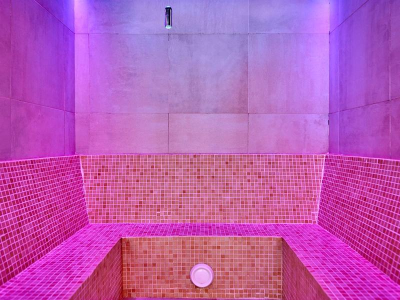 Prázdná parní místnost s růžovými dlaždicemi a fialovým osvětlením.