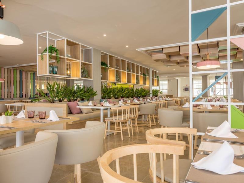 Světá moderní restaurace s dřevěným a bílým nábytkem a zelenými rostlinami.