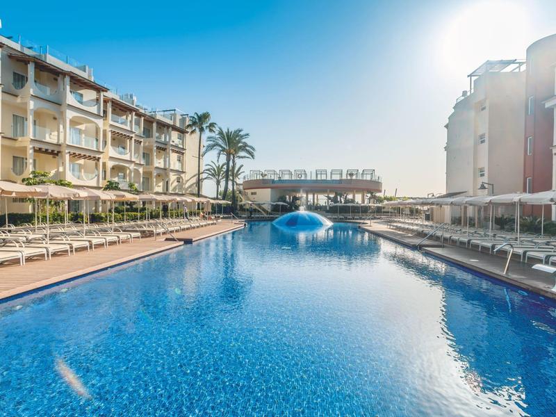 Velký venkovní bazén mezi hotelovými budovami za slunečného počasí.