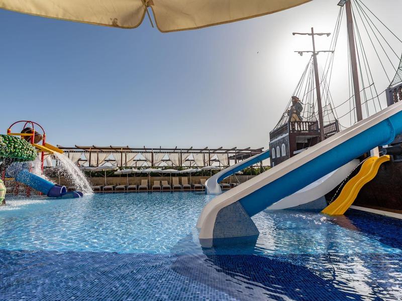 Dětská vodní skluzavka v bazénu se slunečnou oblohou a lodními dekoracemi.