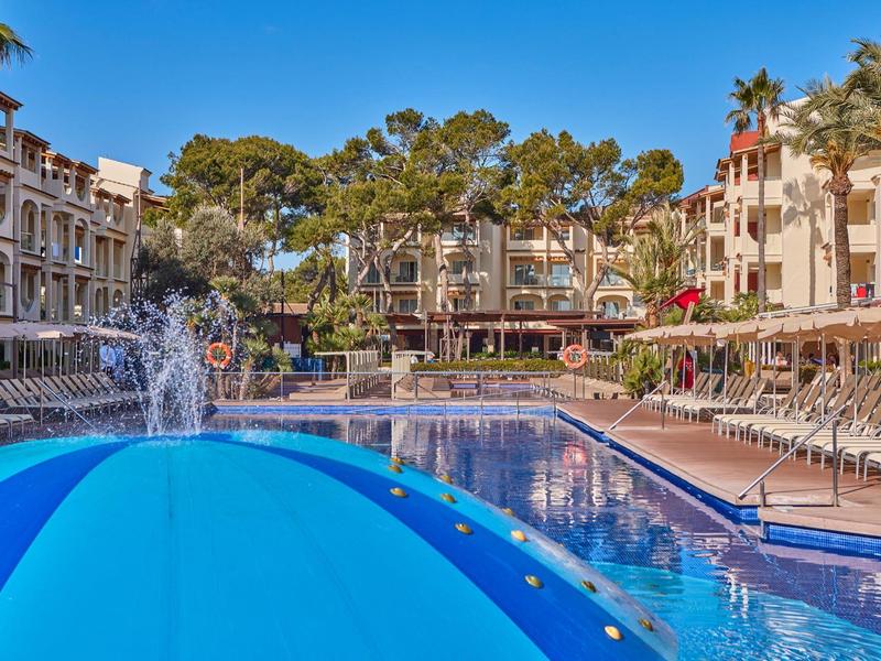 Velký hotelový bazén s modrým slunečníkem a lehátky pod palmami za slunečného počasí.