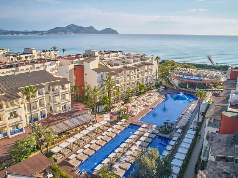 Velké hotelové středisko s několika budovami a velkým bazénem u moře pod jasnou oblohou.