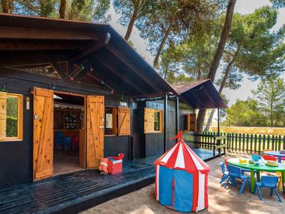 Holzhaus mit offenen Türen neben buntem Kinderzelt und Spieltischen im Freien unter Bäumen