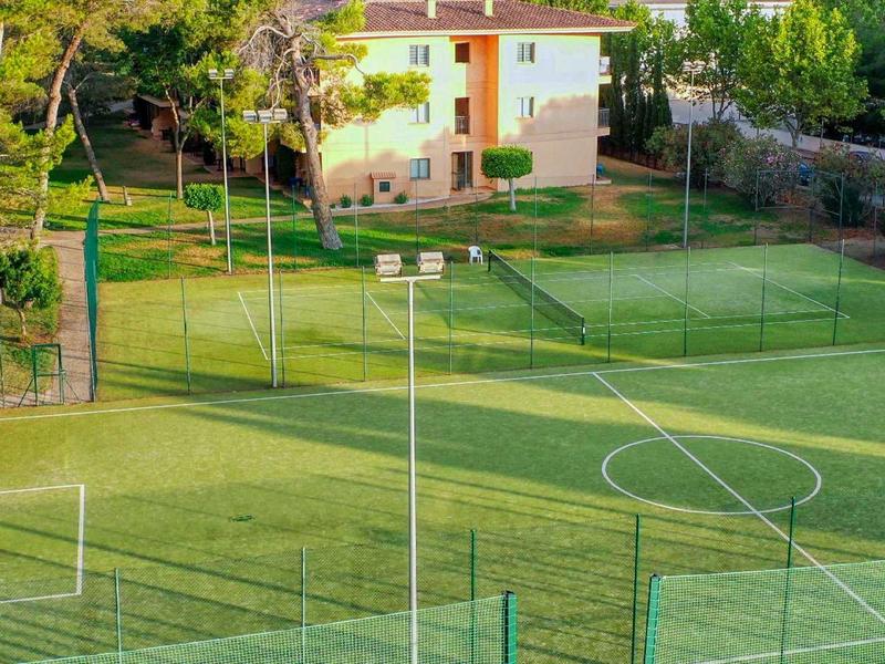 Campo deportivo verde con porterías de fútbol y farolas, rodeado de árboles y un edificio al fondo.