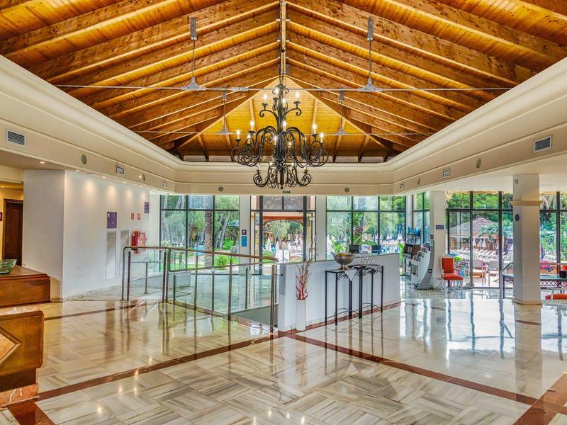 Vestíbulo de hotel luminoso con techo de madera, candelabro y paredes de vidrio con vista al jardín.