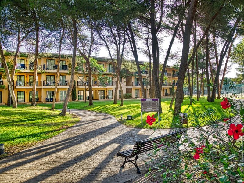 Jardín con bancos, flores rojas y edificio de hotel amarillo entre árboles altos bajo luz solar.
