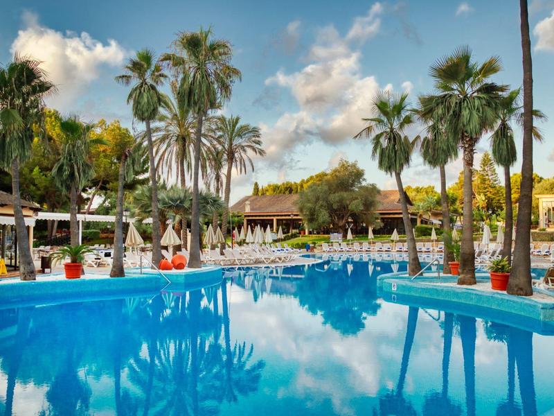 Piscina azul rodeada de palmeras y sombrillas en un complejo hotelero tropical.