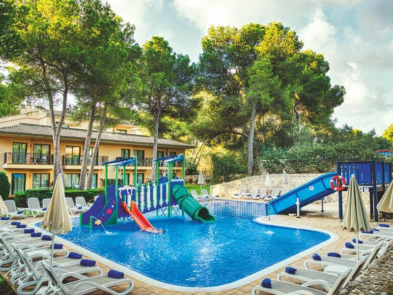 Piscina del hotel con dos toboganes, rodeada de tumbonas y sombrillas grandes bajo árboles.