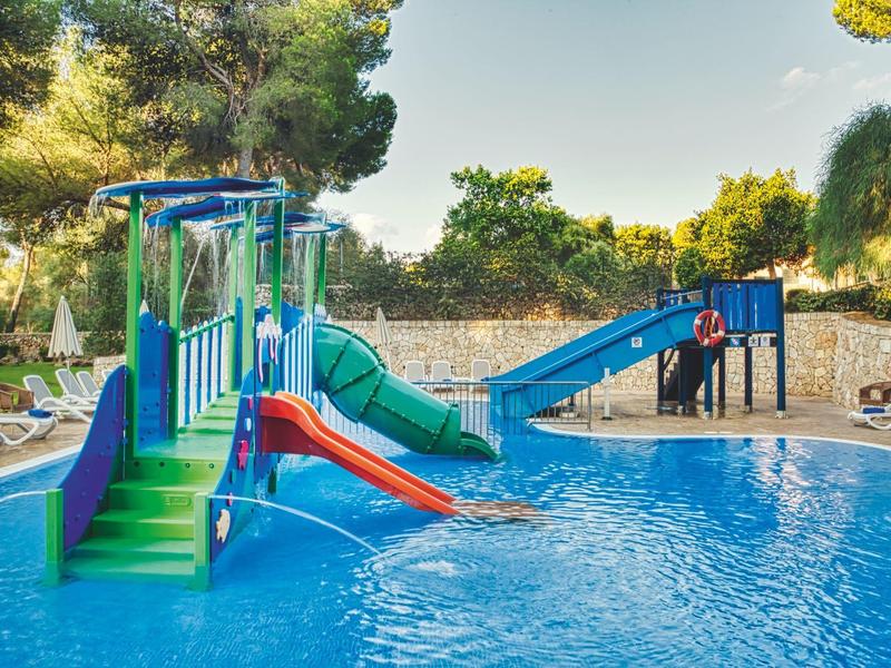 Parque acuático infantil con toboganes en una piscina, rodeado de árboles y muro de piedra.