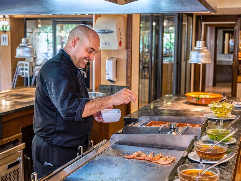 Hombre cocina camarones a la parrilla en una cocina moderna de hotel con diversas salsas e ingredientes.