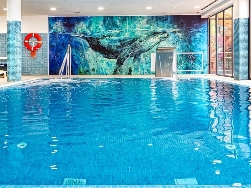 Piscina interior con azulejos azules y mural de ballena en la pared, con ventanas grandes.