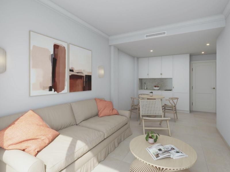 Habitación de hotel luminosa con sofá gris, mesa de centro redonda y cocina en el fondo.