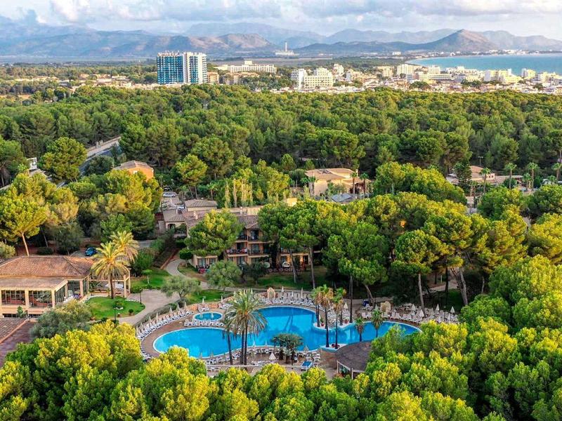 Vista de una piscina de hotel rodeada de árboles verdes con montañas al fondo bajo un cielo despejado.