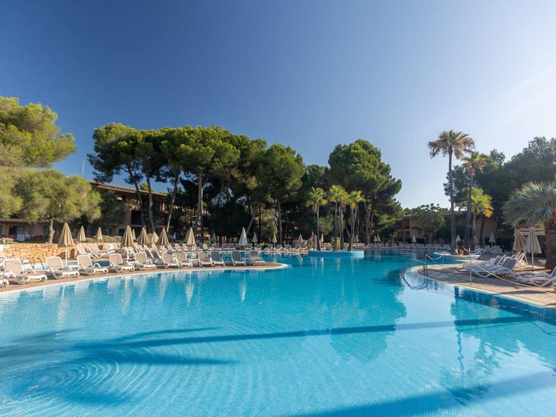 Gran piscina azul con tumbonas y sombrillas, rodeada de árboles y cielo despejado.