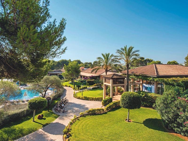 Complejo hotelero con jardín, piscina, palmeras y senderos bajo un cielo despejado en un día soleado.
