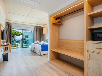 Helles Hotelzimmer mit Holzschrank, Doppelbett und Balkon mit Blick auf Wasser und Bäume.