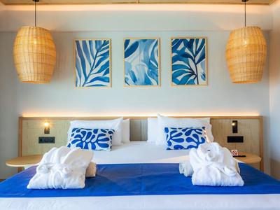Modernes Schlafzimmer mit großem Bett, blauen Kissen, weißen Bademänteln und Wandbildern mit Blattmotiven.