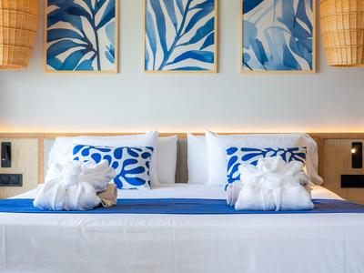 Modernes Schlafzimmer mit großem Bett, blauen Kissen, weißen Bädern und dekorativen Pflanzenbildern.