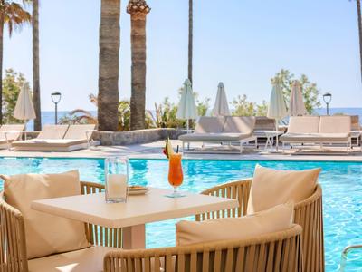 Moderner Außenbereich mit Holztisch, Sesseln, blauen Cocktails und Pool mit Palmen im Hintergrund.