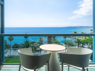 Balkon mit zwei grauen Stühlen und rundem Tisch, Blick auf Meer, Palmen und blauen Himmel.