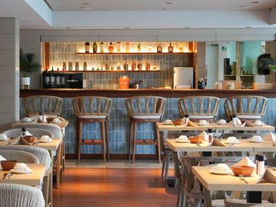 Modern eingerichtetes Restaurant mit Holzstühlen, gedeckten Tischen und beleuchtetem Barbereich.