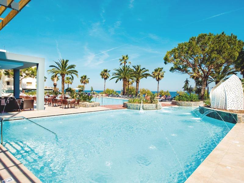 Duży basen na świeżym powietrzu z tarasem i palmami, widok na morze pod błękitnym niebem.