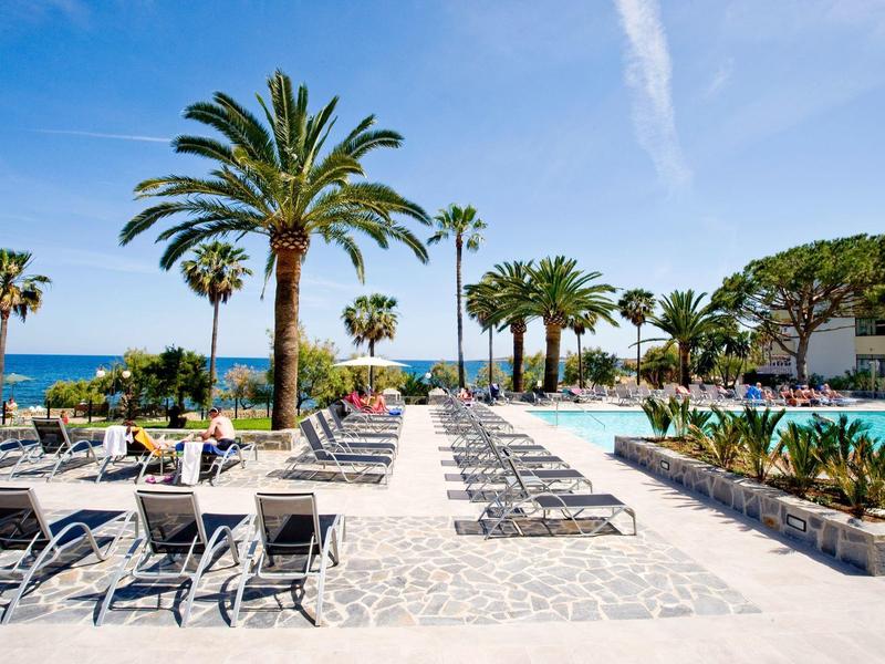 Strefa basenu hotelowego z leżakami i palmami pod błękitnym niebem z widokiem na morze.