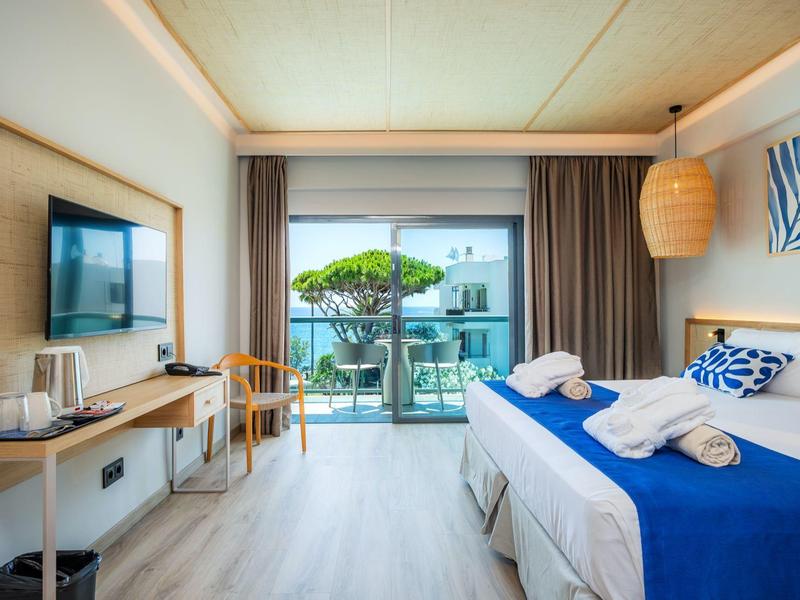 Nowoczesny pokój hotelowy z łóżkiem podwójnym, TV, biurkiem i balkonem z widokiem na basen i morze.