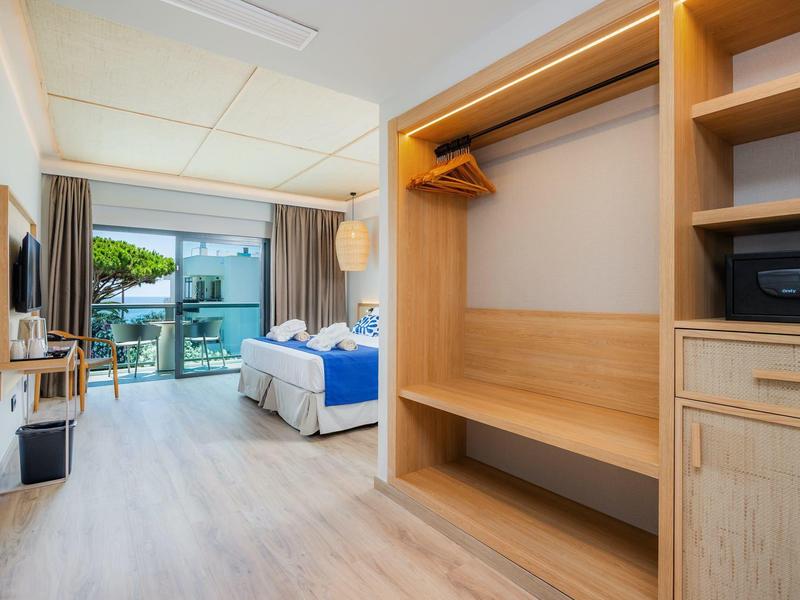 Jasny pokój hotelowy z otwartą szafą, podwójnym łóżkiem i balkonem z widokiem na ogród.