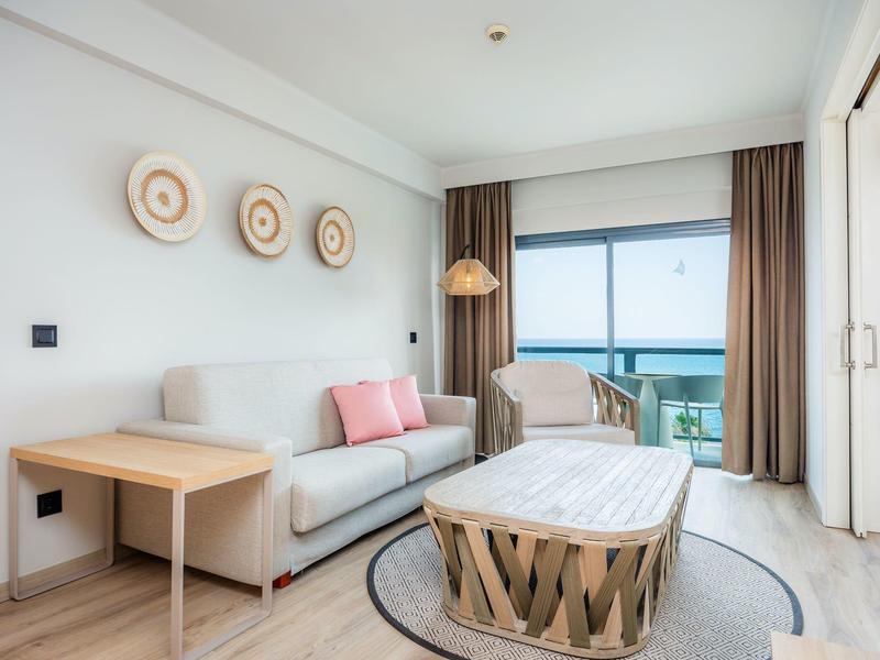 Jasny salon z sofą, stołem i widokiem na morze przez duże okno balkonowe.