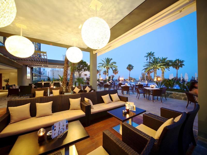 Przytulny taras hotelowy z wygodnymi miejscami do siedzenia z widokiem na oświetlony basen i palmy o zmierzchu.