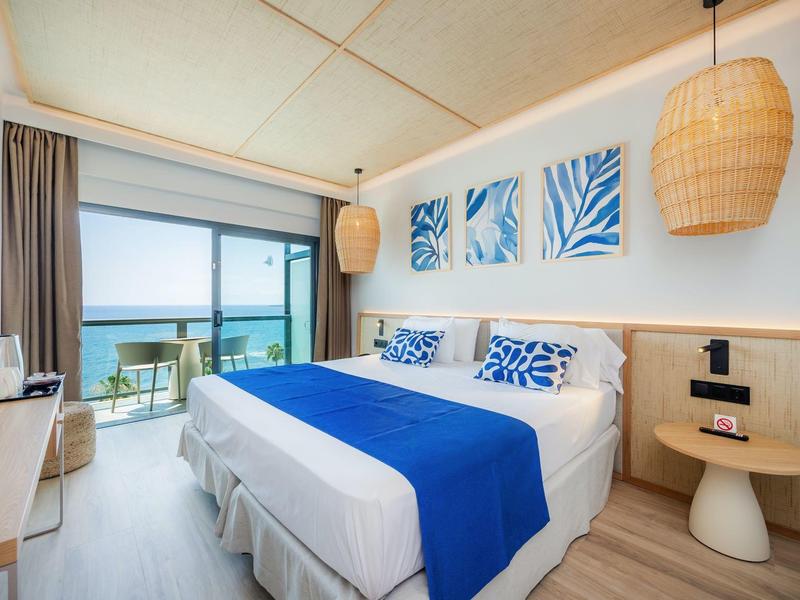 Jasny pokój hotelowy z łóżkiem podwójnym, niebiesko-białą dekoracją i widokiem na morze z balkonu.