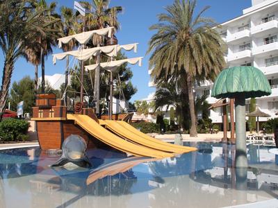 Strefa basenowa z zjeżdżalnią w kształcie statku pirackiego, palmami i hotelem w tle.