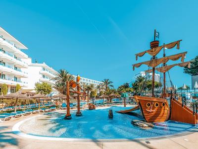 Zwembadgedeelte bij resort met palmbomen, ligstoelen en een piratenschip-speelstructuur onder blauwe lucht.